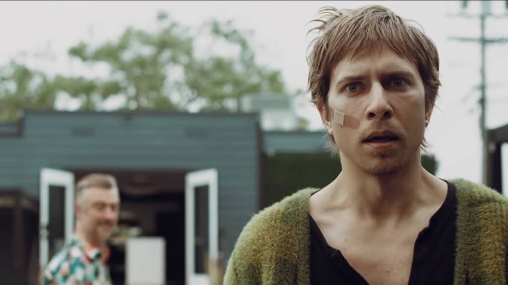 Aaron Holliday in the foreground has a bandaid on his cheek in character as Duke as Sean Gunn as Clint Binto stares at Duke in the blurred background.