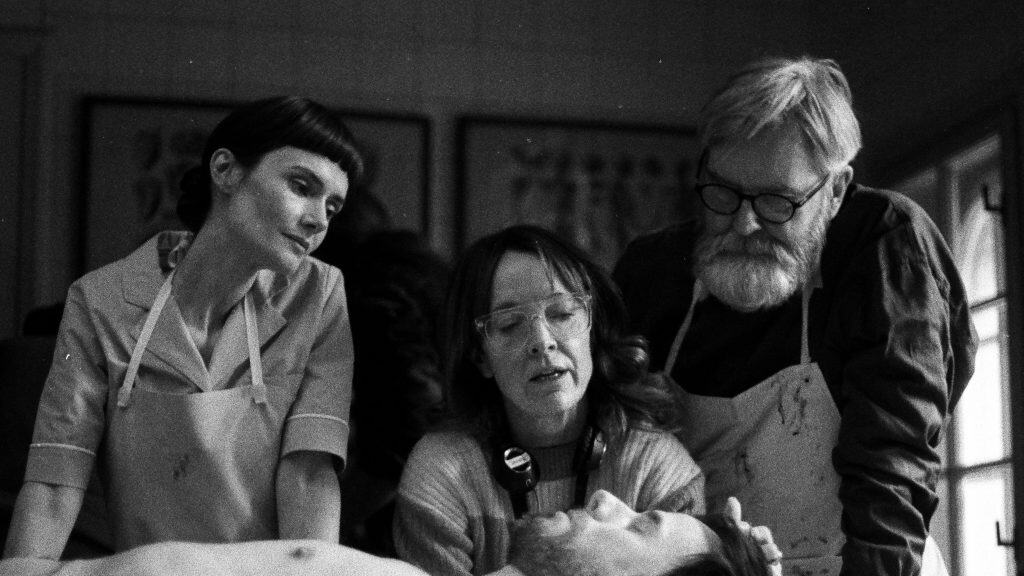 Three people in aprons examine a body on a table