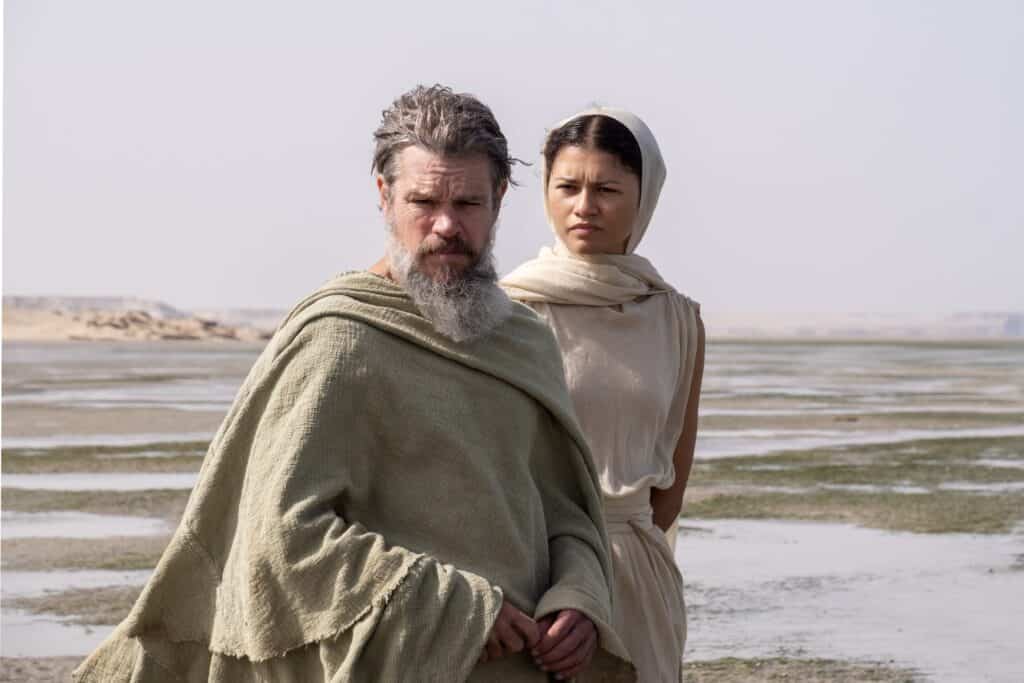 Matt Damon as older Odysseus and Zendaya as Athena, walking just behind him on Ogygia's beach.