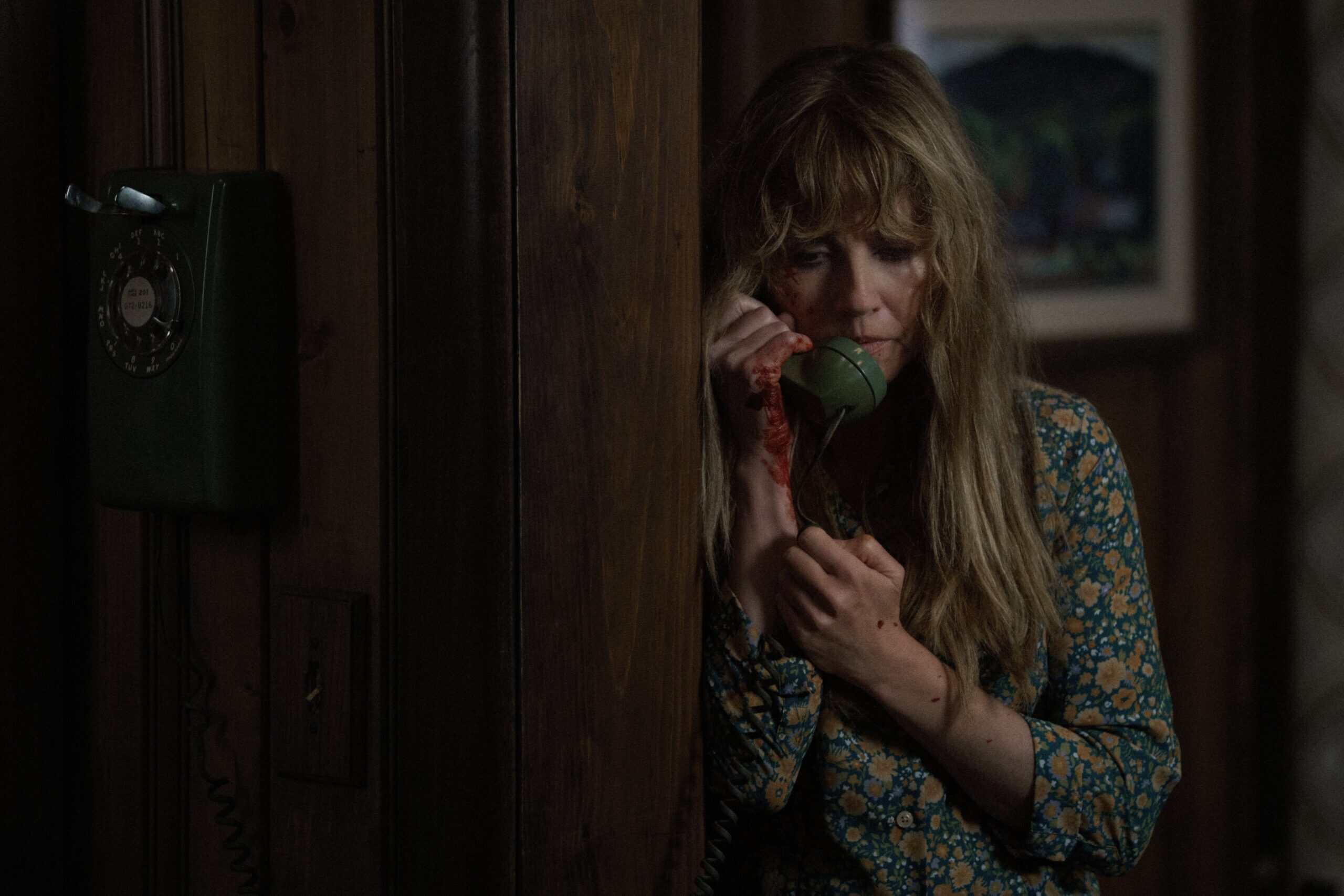 Linda Cardellini as Pamela Voorhees. She's holding a phone receiver as she leans against the wall, talking to someone on the other end of the line. Her hands are covered in blood.