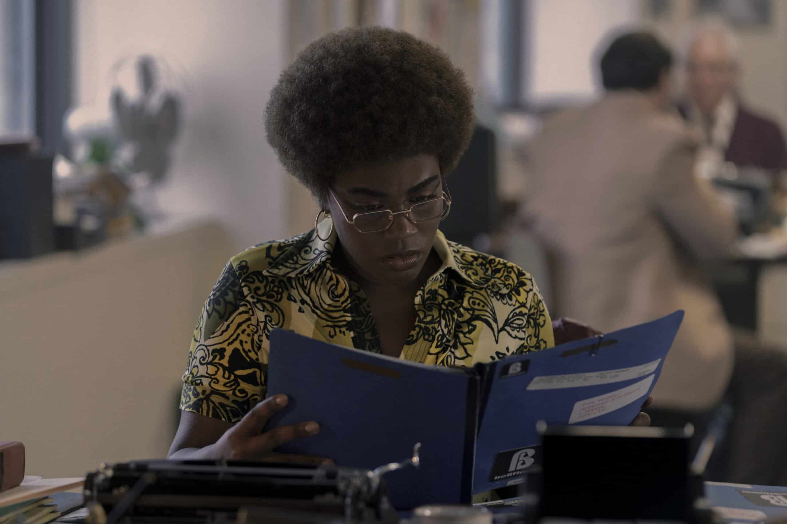 Devin Kessler as Brianna. She sits at a desk as she reads attentively documents included in a blue folder.