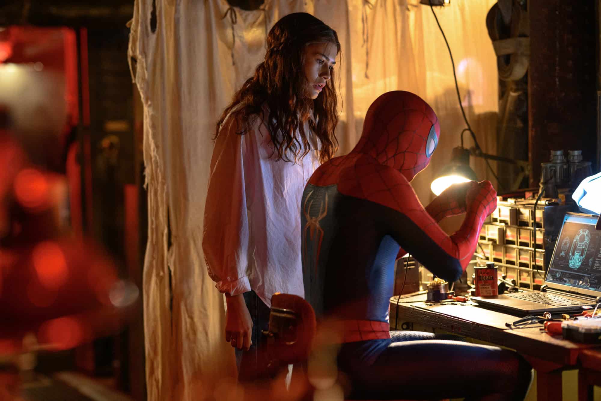 Zendaya as MJ and Tom Holland as Peter Parker/Spider-Man in a scene of Spider-Man: Brand New Day. Peter wears his Spider-Man suit and works at a desk while MJ stands beside him and looks over at an object Peter holds in his hands.