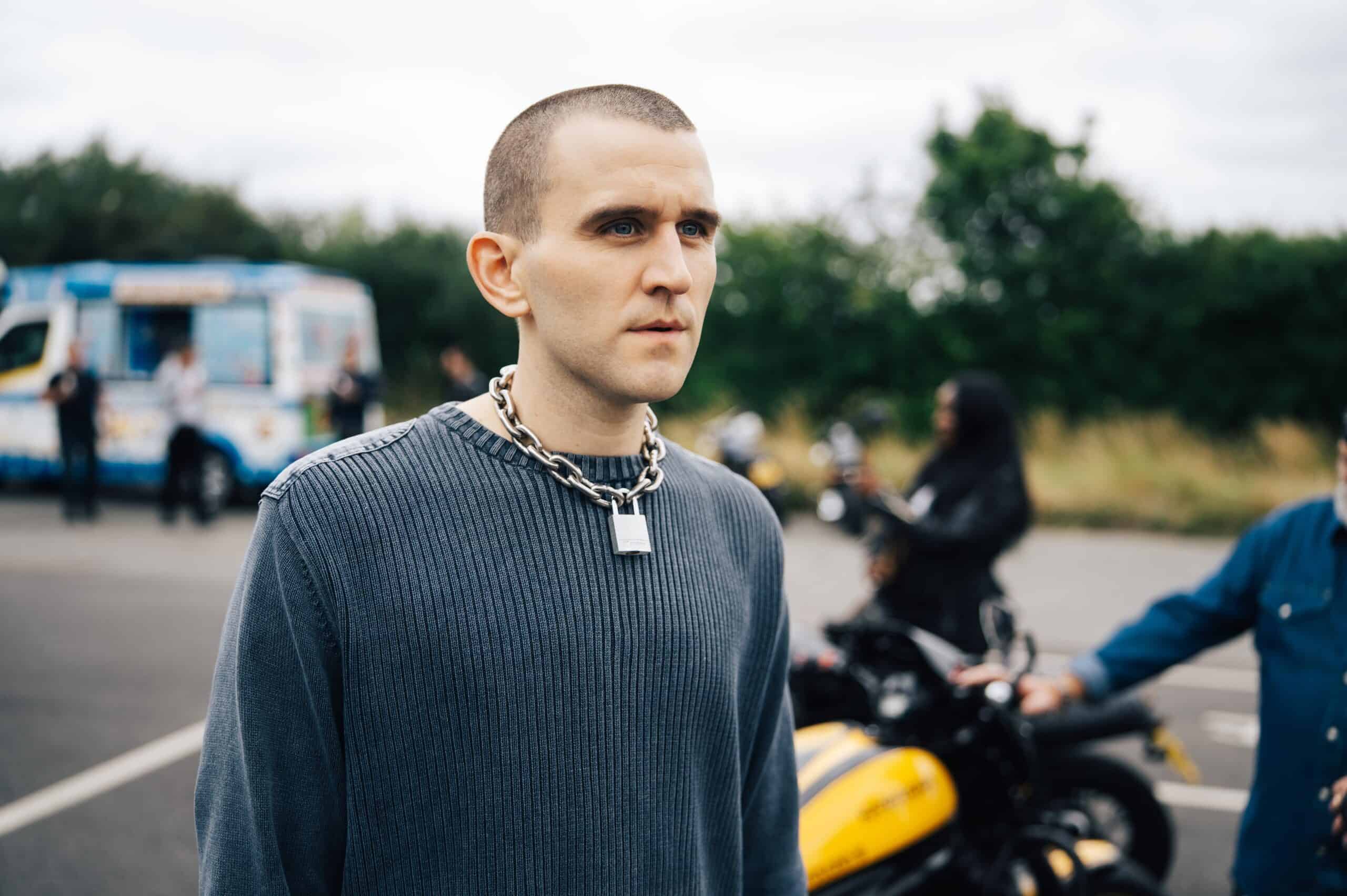 Harry Melling as Colin in a scene of Pillion. He's wearing a blue-grey sweatshirt with a neck chain and a padlock around his neck and rocks a buzzcut. In the background, there are people on their motorcycle in a parking lot.
