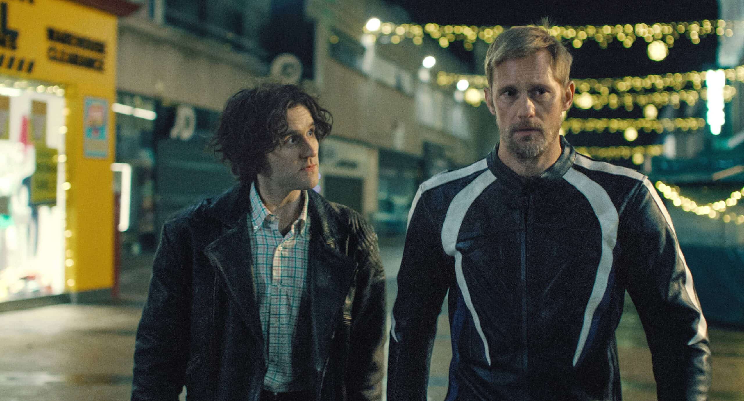 Harry Melling as Colin and Alexander Skarsgård as Ray in a scene of Pillion. The pair walks around a deserted Bromley street. as Christmas lights can be seen in the background.